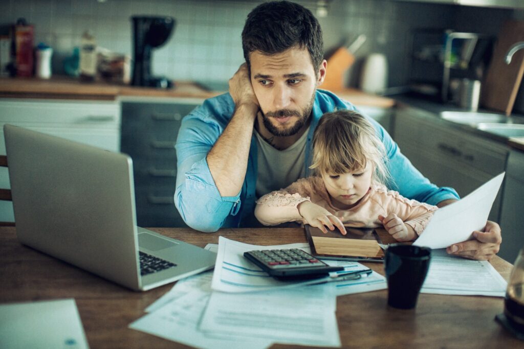 mies stressaantuneena istuu keittiön pöydän äärellä pieni lapsi sylissään, mies katsoo tietokoneen ruutua ja pöydällä on papereita
