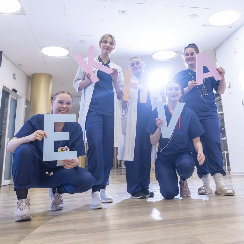Hoitajat ja lääkärit poseeraavat iloisina sairaalan käytävällä EKHVA -kirjainten kanssa.